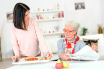 Frau sitzt mit Zeitung an Tisch, andere Frau stellt Tablett mit Essen neben sie.